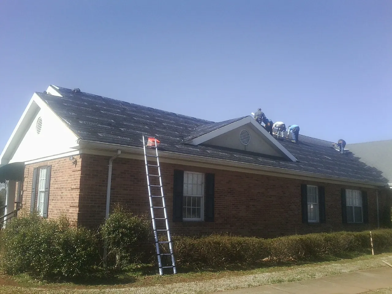 Crew tearing shingles off a brick ranch roof with a ladder set — Metal Roof Repair work by our crew in Collinston