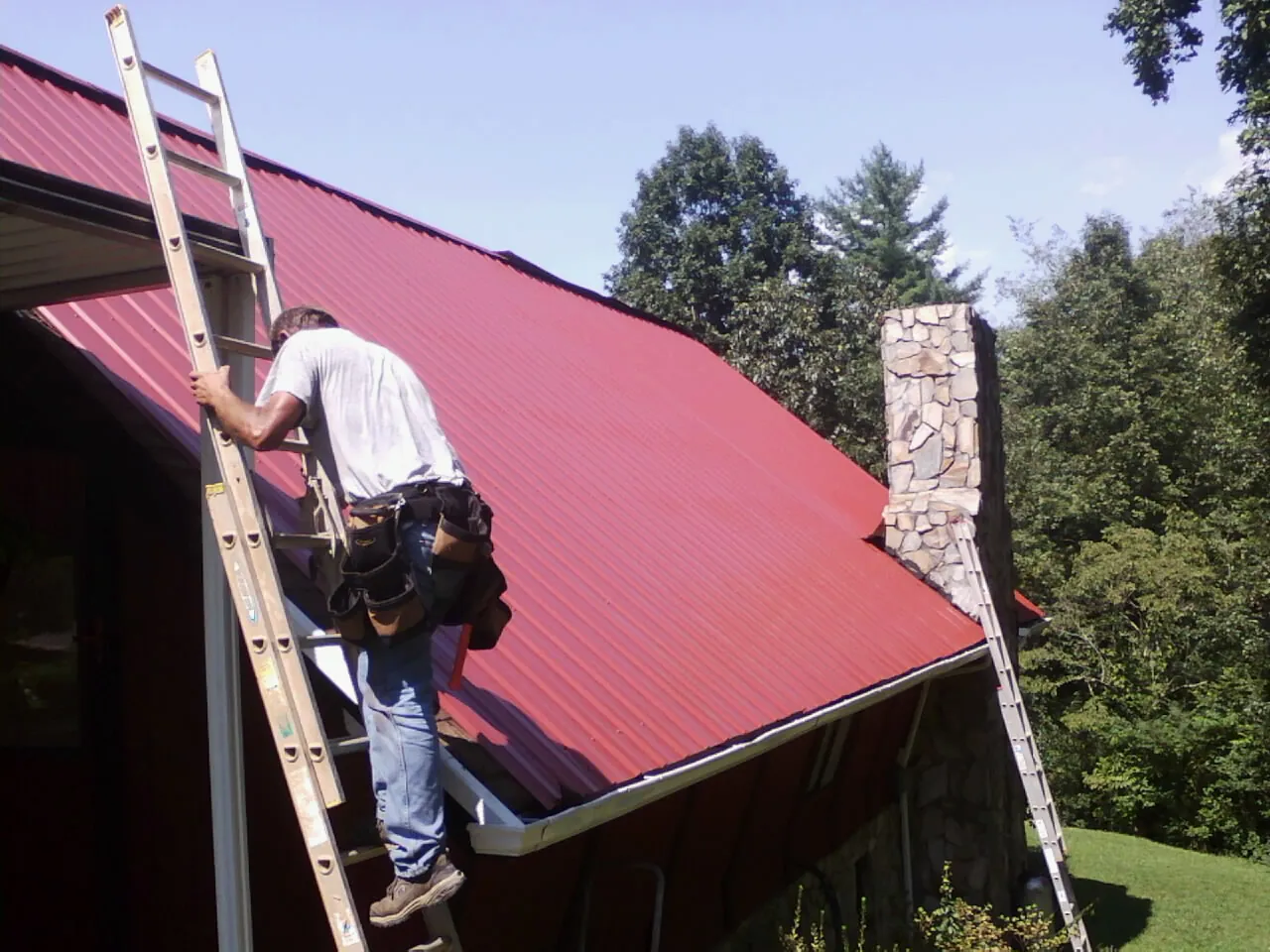 Roofer on a ladder beside a red metal roof and stone chimney — Soffit & Fascia Repair by Roofing Brothers in Collinston